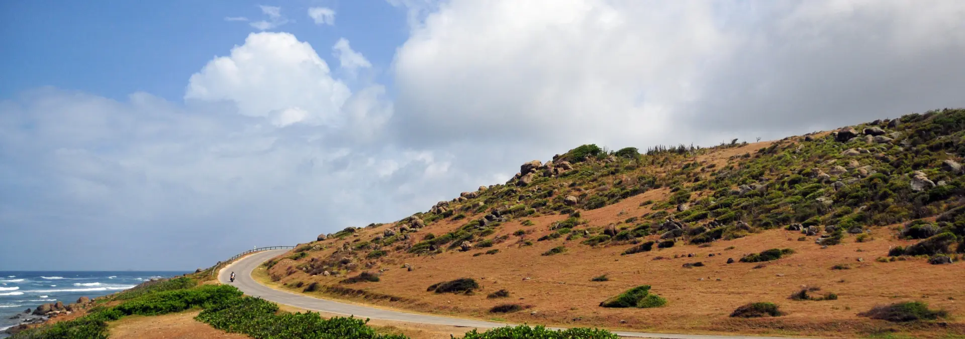 Scenic view of Coralita Road along the rocky northeast coast of St. Martin, with ocean waves and grassy hillside.