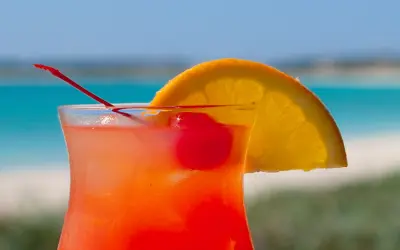 Bright red-orange rum punch cocktail garnished with cherries and an orange slice, by the sea.