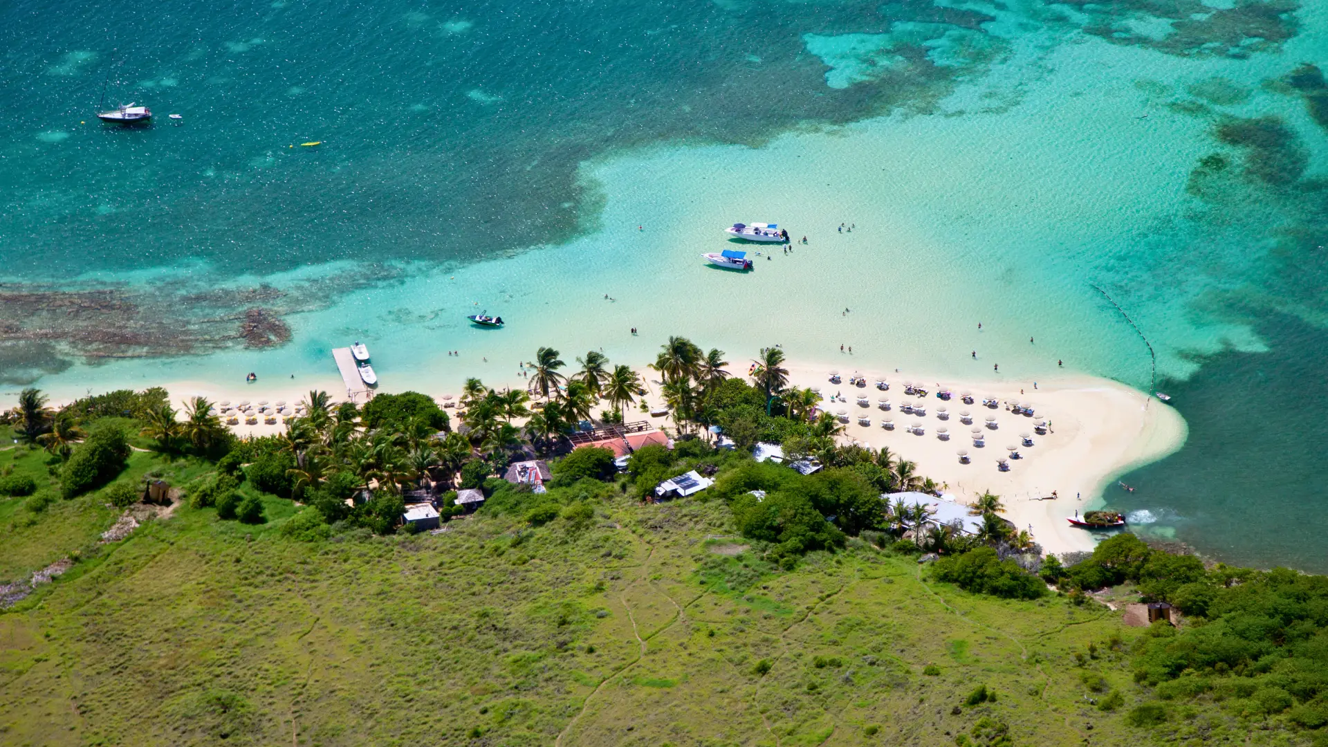 Aerial view of Pinel Island, St. Martin, highlighting the crystal-clear water, boats, and beach umbrellas along the coastline. Discover more with your Caribbean Concierge