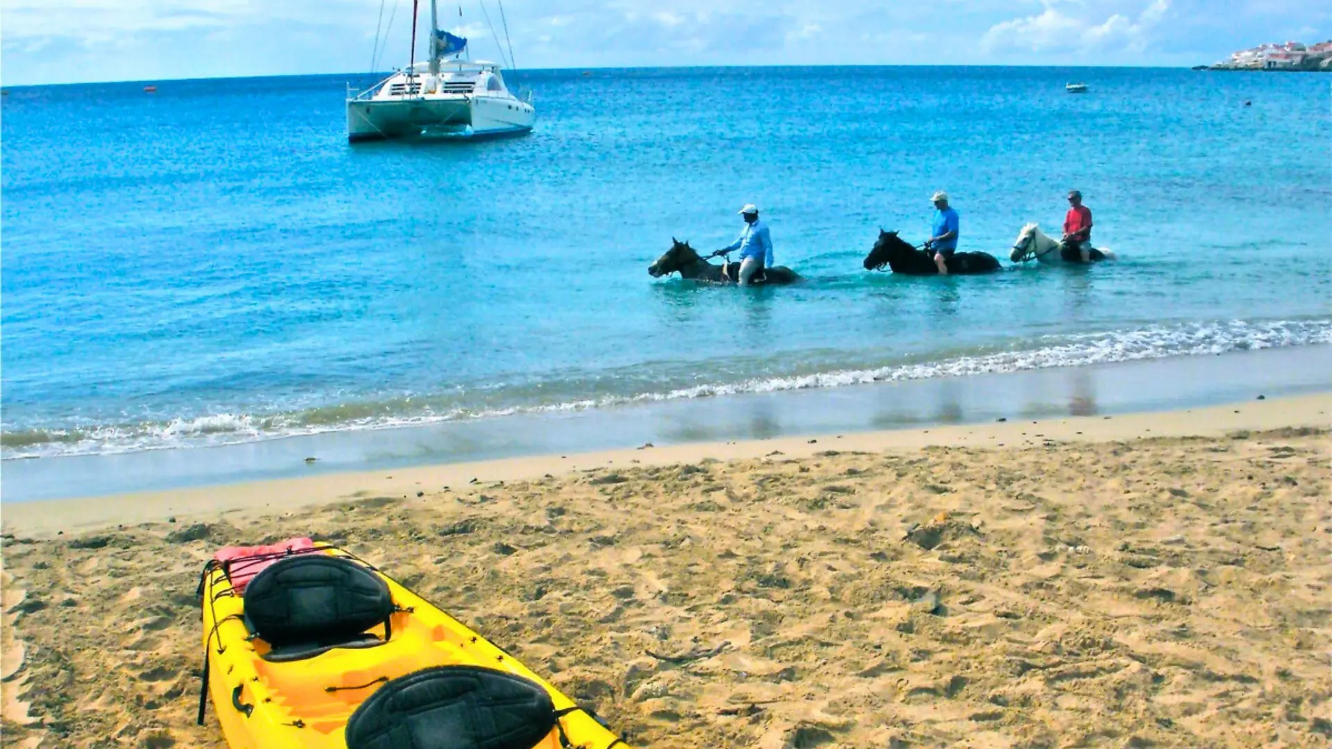 Riders on horseback entering the Caribbean Sea beside a yellow kayak on a sandy beach in St Maarten