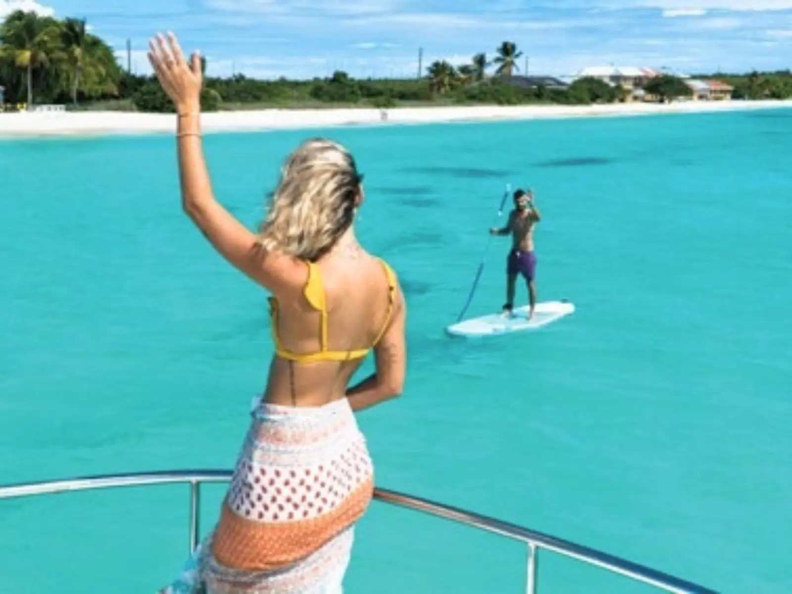Woman standing on boat deck waving toward a paddleboarder in shallow turquoise water
