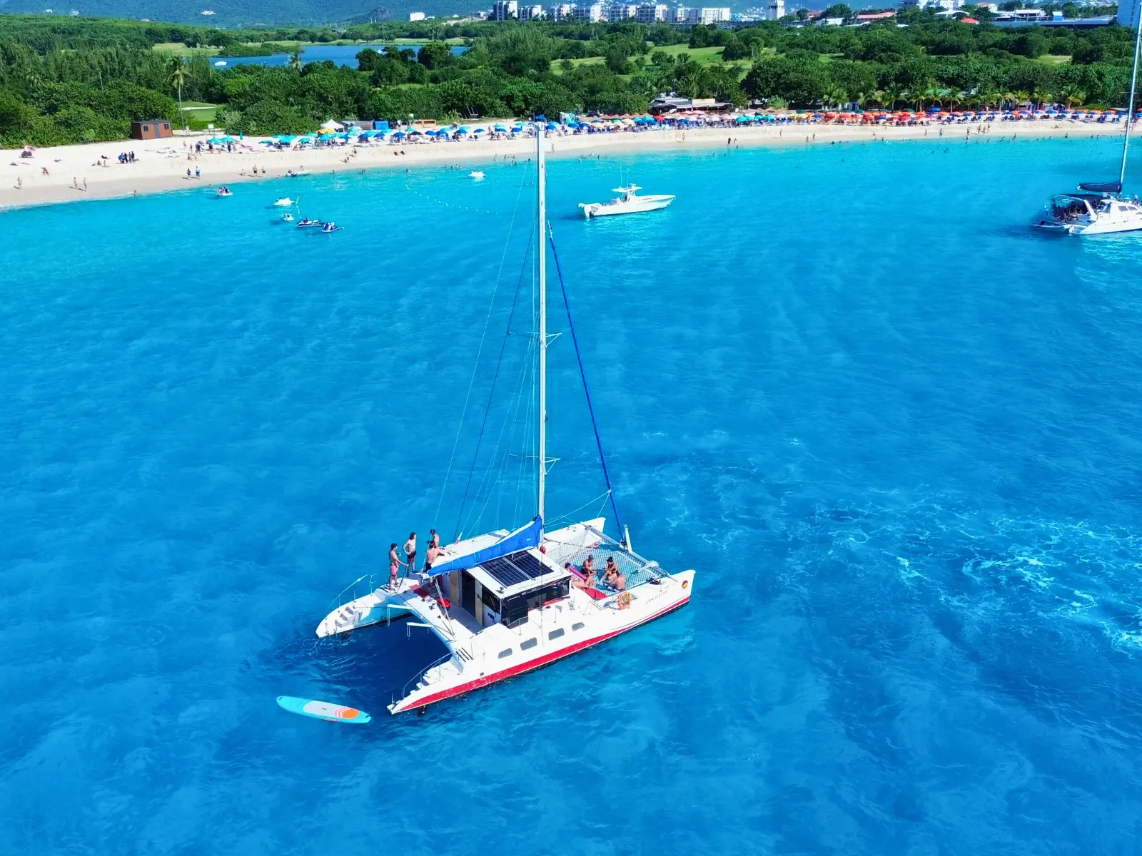 Drone view of a private catamaran anchored off a busy St Maarten beach with umbrellas, swimmers, and boats nearby