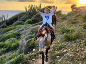 Smiling child riding a horse along a scenic coastal trail in St Maarten with sunset light and ocean views