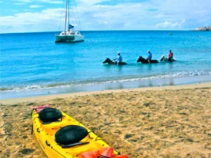 Group of riders on horseback walking through calm coastal waters with a catamaran anchored offshore in St Maarten